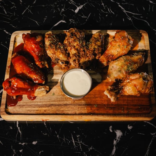 A wooden board with three types of chicken wings and a small cup of dipping sauce.