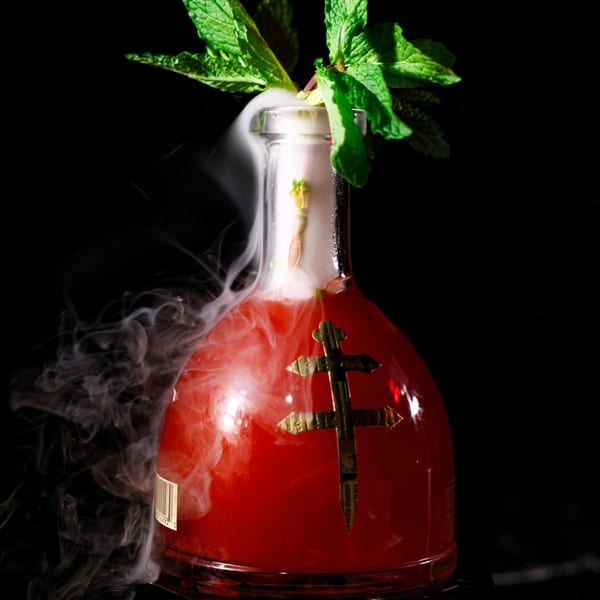 A red cocktail in a decorative bottle with a gold cross, garnished with mint and emitting smoke.
