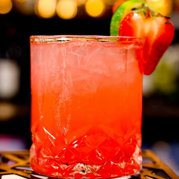 A vibrant red cocktail in a textured glass, garnished with a fresh strawberry and lime.