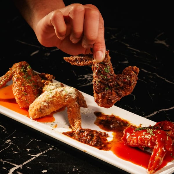 A hand holds a saucy chicken wing above a plate with three different flavored wings.